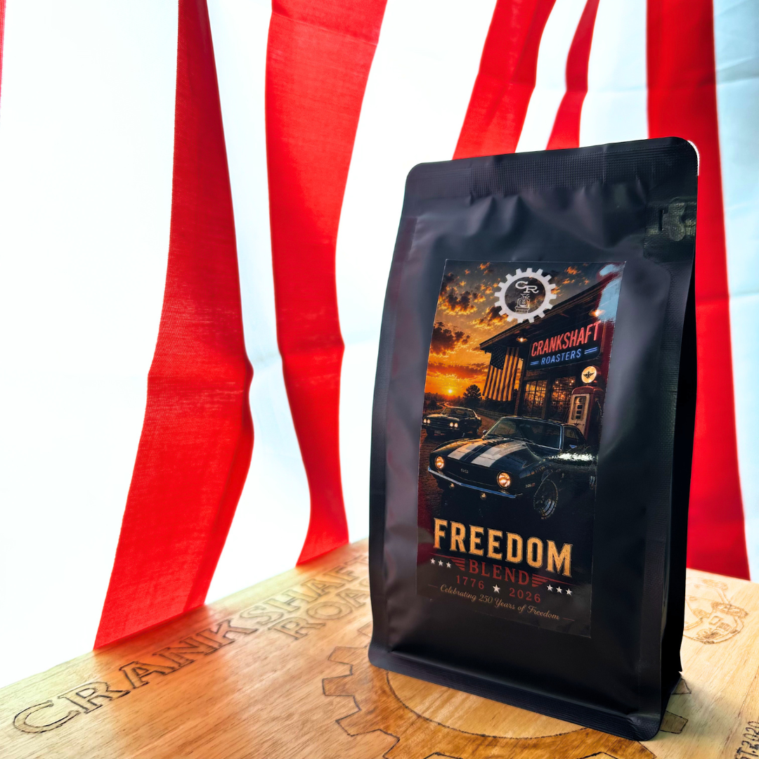 Black bag of Crankshaft Roasters Freedom Blend coffee featuring a patriotic muscle car label, displayed on a wooden surface with a large American flag backdrop.